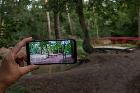 woman making a photo with smartphone of the typical red japanese bridge in garden with forest trees and plants and sand at the border of small water.の写真素材