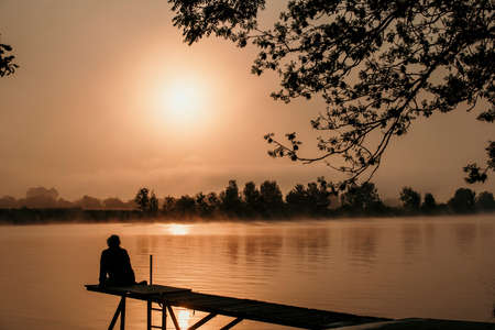 silhouette woman at wooden jetty during sunset in the early morning over the river maas in limburg...