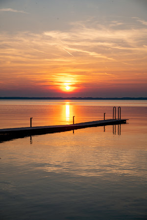 the sun sets over the water with trees in the foreground a wooden decking on the left of the photoの写真素材