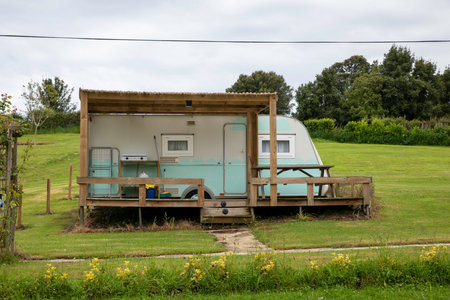 Charming vintage caravan with sheltered deck set in open green meadow surrounded by trees. Perfectly encapsulates serene getaway ambiance for nature lovers seeking tranquility and rustic accommodation optionsの写真素材