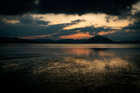 A tranquil lake at dusk with dark clouds and a soft orange glow along the horizon. Distant hills...