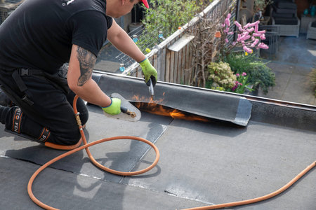 rotterdam,holland,23-march-2026:Roofer uses a propane torch to heat and torch-on waterproofing membrane on a flat residential roof during repair and maintenance, showing professional roofing work, safety gloves, and outdoor construction activity.のeditorial素材
