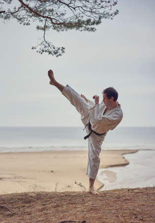 Karate man in an old kimono and black belt training high kick at the sea. Martial arts concept. The river flows into the sea at the background.の写真素材