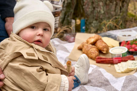 Small baby in the jacket and white cap with funny ears holding food in the hand on the picnic at the forest. She looking at the camera. Miscellaneous food at the background.の写真素材