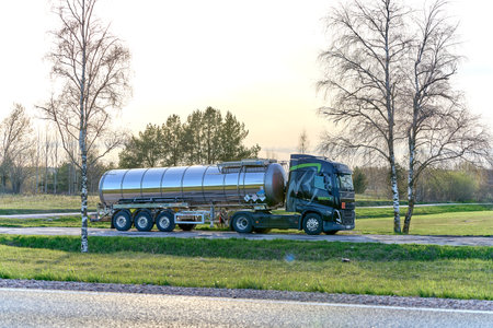 Big metal fuel tanker truck shipping fuel standing on the countryside road near the green field and birch without leafs.のeditorial素材