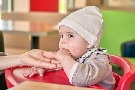 A little brown eyed girl with big lashes in a light cap sitting in a red childs chair eating a fruit. Mother hold hand to help.の写真素材