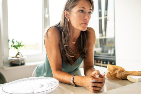 A woman leans casually on a kitchen counter, embodying a modern lifestyle that fosters reflection and tranquilityの写真素材