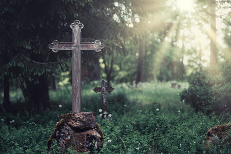 A tranquil graveyard with a weathered metal cross stands on a stone, surrounded by stunning nature and warm sunlight filtering throughの写真素材