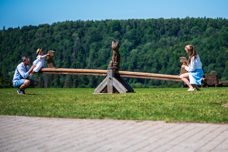 Beautiful caucasian family is having fun on playground. Parents with children riding on a swing. Sunny summer day. Green forest at the background.の写真素材
