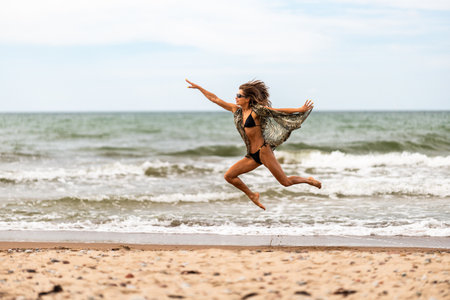 A vibrant beach scene capturing a carefree woman joyfully leaping high over the warm sand and gentle wavesの写真素材