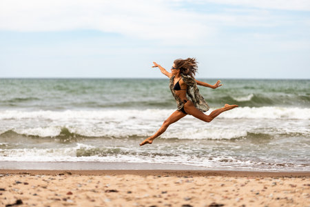 A lively and energetic woman joyfully jumps on a beautiful sandy beach, embodying freedom against ocean wavesの写真素材