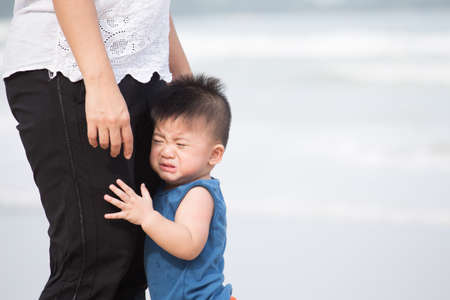 Asian baby boy is standing, fearing and hugging mother legs while visiting the sea for the first...