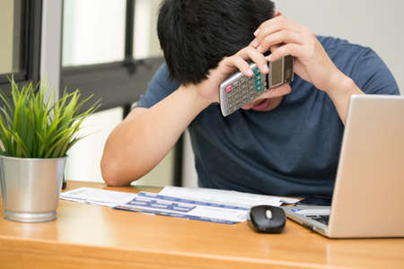 Asian man stressed while calculate the credit card debt with a calculator and working on laptop at home, Sad and dramatic,Â  Debt stress and mental health conceptの写真素材