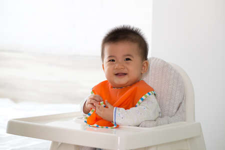 Cheerful happy Asian baby waiting to eat food and sitting on child chairの写真素材