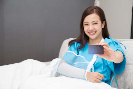 Asian woman holding insurance cards and wear patient suits on the arms wearing soft splint sitting on bed at hospitalの写真素材