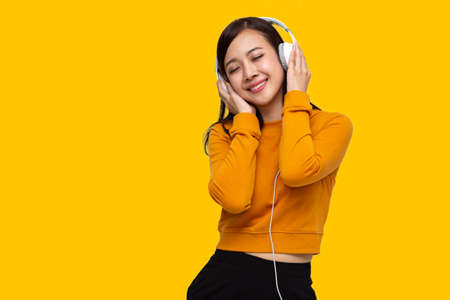 Portrait of happy beautiful young Asian woman listening music on her white headphones, dancing and smiling isolated on yellow backgroundの写真素材