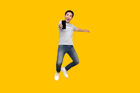 Asian man jumping and presenting mobile phone isolated on yellow background