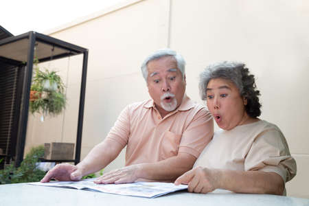 Excited Asian senior mature couple feeling amazed by good surprise news while reading newspaper or...