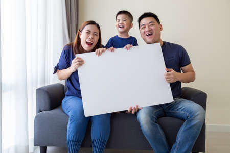 Happy Smiling Asian family holding blank big white poster and sitting on sofa in living room, Wow...