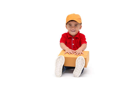Happy kid Asian boy delivery man in yellow cap and red shirt sitting with parcel post box isolated...
