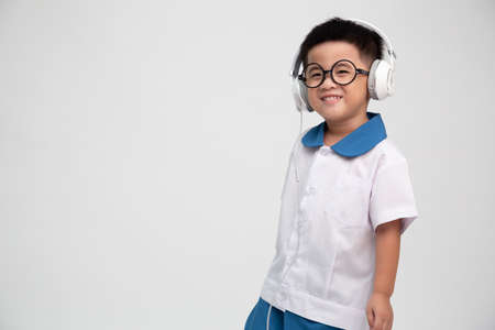 Cheerful smiling asian little boy in student uniform listening music with headphone isolated on...
