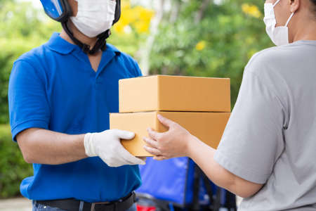 Asian delivery man wearing face mask and blue uniform with motorcycle delivering parcel box express...