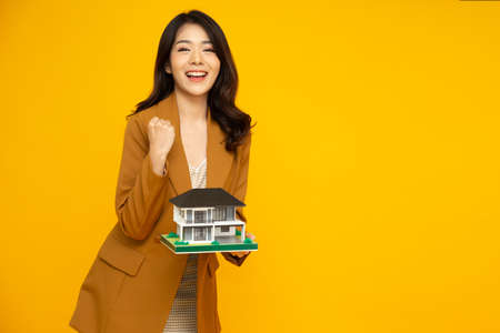 Asian businesswoman smiling and holding house sample model isolated on yellow background, Real estate and home insurance conceptの写真素材
