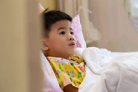 Asian little boy wearing patient outfits lying on the bed in hospital, Worried expression concept