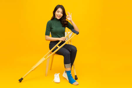 Young Asian woman sitting on chair and sprain foot using crutches and showing ok sign isolated on yellow background, Personal accident conceptの写真素材