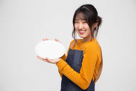 Young Asian woman in apron standing and holding empty white plate or dish isolated on white backgroundの写真素材
