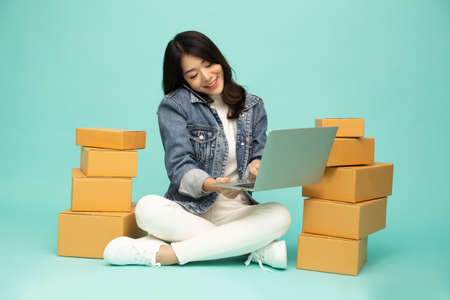 Happy young Asian woman small business talking on the phone and sitting on floor with parcel box and...