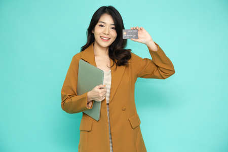 Young beautiful Asian businesswoman in suit holding computer laptop and smiling, showing, presenting credit card for making payment or paying online business isolated on green backgroundの写真素材
