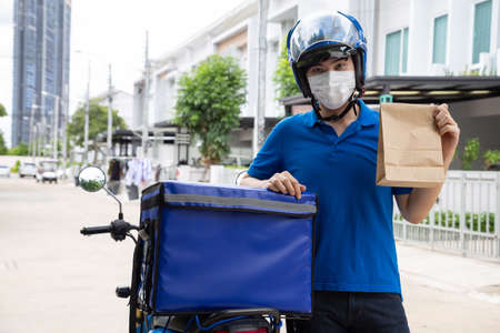Young Asian man wearing face mask and holding food paper bag and standing on the side of the delivery box, Motorcycle delivering food express service conceptの写真素材