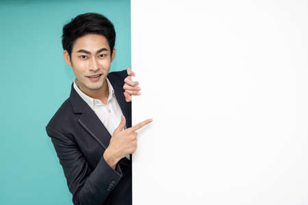 Young asian man in suit standing behind blank white billboard and pointing with finger to empty copy spaceの写真素材