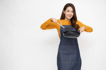 Young Asian woman housewife wearing kitchen apron cooking and holding pan and spatula isolated on white backgroundの写真素材
