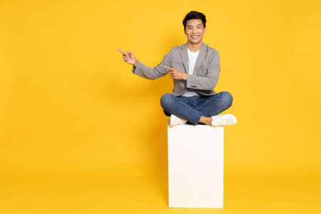 Smiling young Asian businessman pointing to empty copy space and sitting on white box isolated on yellow backgroundの写真素材