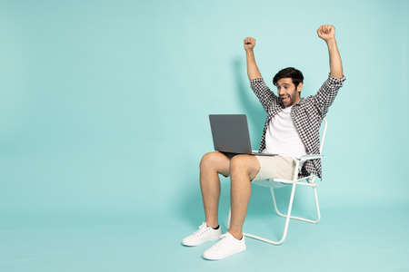 Man using laptop computer and sitting on canvas chair and hands up raised arms from happiness isolated on green background, Excited man winner success in holiday conceptの写真素材