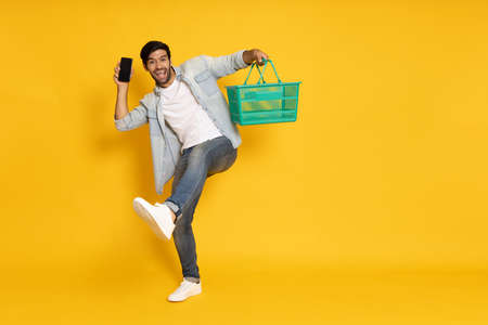 Happy man having fun and holding shopping basket and mobile phone isolated on yellow studio backgroundの写真素材