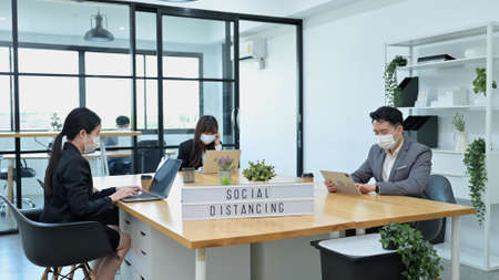 Group of young Asian team business people with face masks work in office and keep distancing in company workplace, Social Distancing conceptの写真素材