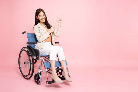 Smiling young Asian businesswoman sitting on wheelchair and pointing to empty copy space isolated on pink backgroundの写真素材