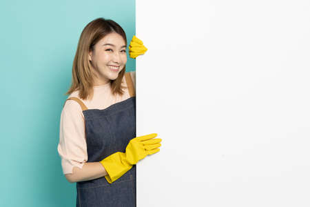 Portrait of Happy Young Asian woman maid or housewife wearing rubber gloves is standing behind the white blank banner or empty copy space advertisement board on green backgroundの写真素材