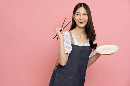 Young Asian woman with jeans apron holding chopsticks and empty white plate or dish isolated on pink...