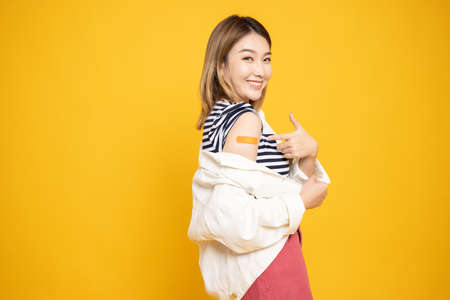 Young Asian woman showing arm with plaster or bandage of Covid-19 Vaccinated isolated on yellow backgroundの写真素材