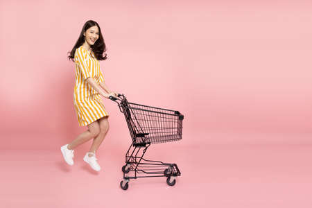 Full length portrait of young Asian woman jumping and pushing an empty shopping cart or shopping trolley isolated on pink backgroundの写真素材