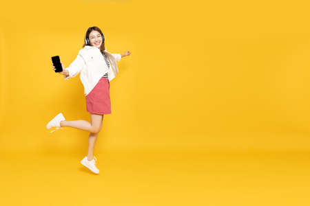 Young Asian woman listening favourite music on mobile phone application with wireless headphones and dancing isolated on yellow background, Full body compositionの写真素材