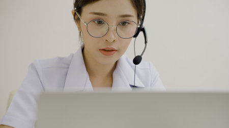Close up of a Asian female doctor wearing a lab coat and headphone sitting in the medical room at hospital looking at a computer screenの写真素材