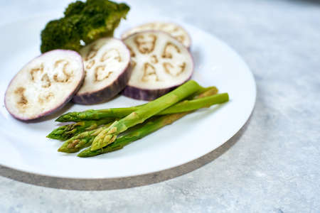 Steamed vegetables in a white plate on a blue table. Eggplant, broccoli, asparagus.の写真素材