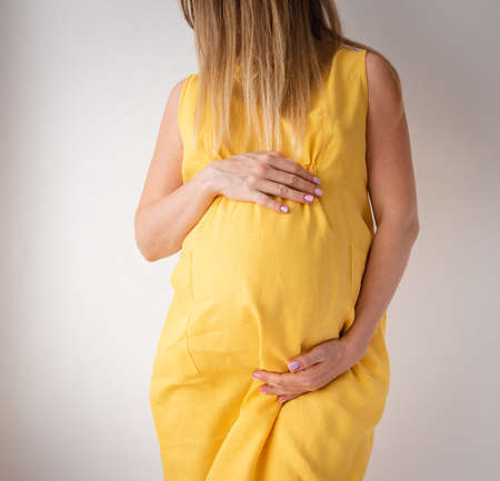 pregnant woman in a yellow dress stands on a white background with hands on her stomach, the concept of femininityの写真素材