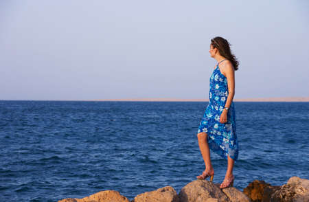 The girl in a blue dress standing on a stone and looking at the seaの写真素材