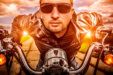Biker man wearing a leather jacket and sunglasses sitting on his motorcycle looking at the sunset. Filter applied in post-production.の写真素材